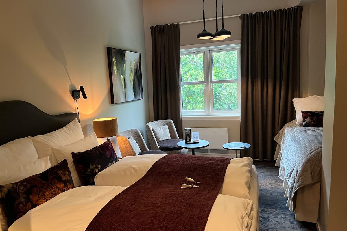 A hotel room with two beds, a small table, a chair, a painting on the wall, and a window with curtains.