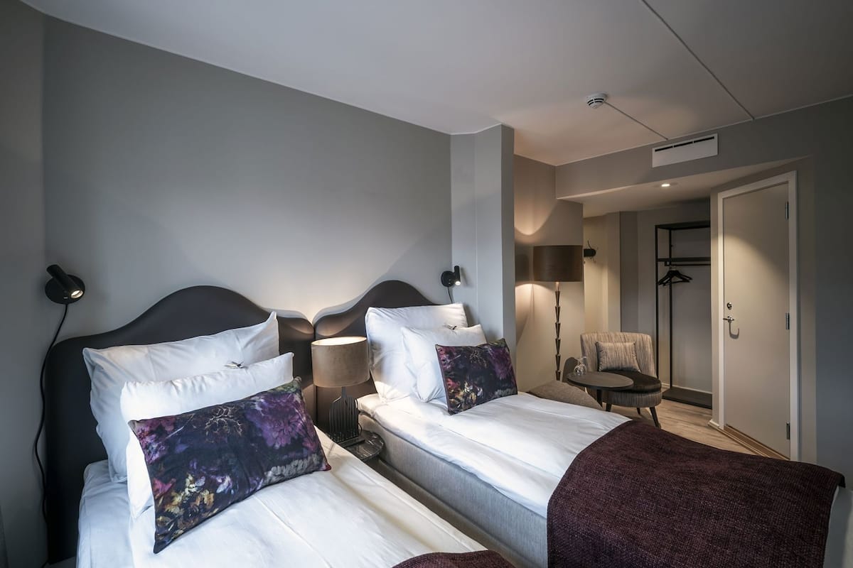 A hotel room with two beds, a chair, a lamp, and a door.