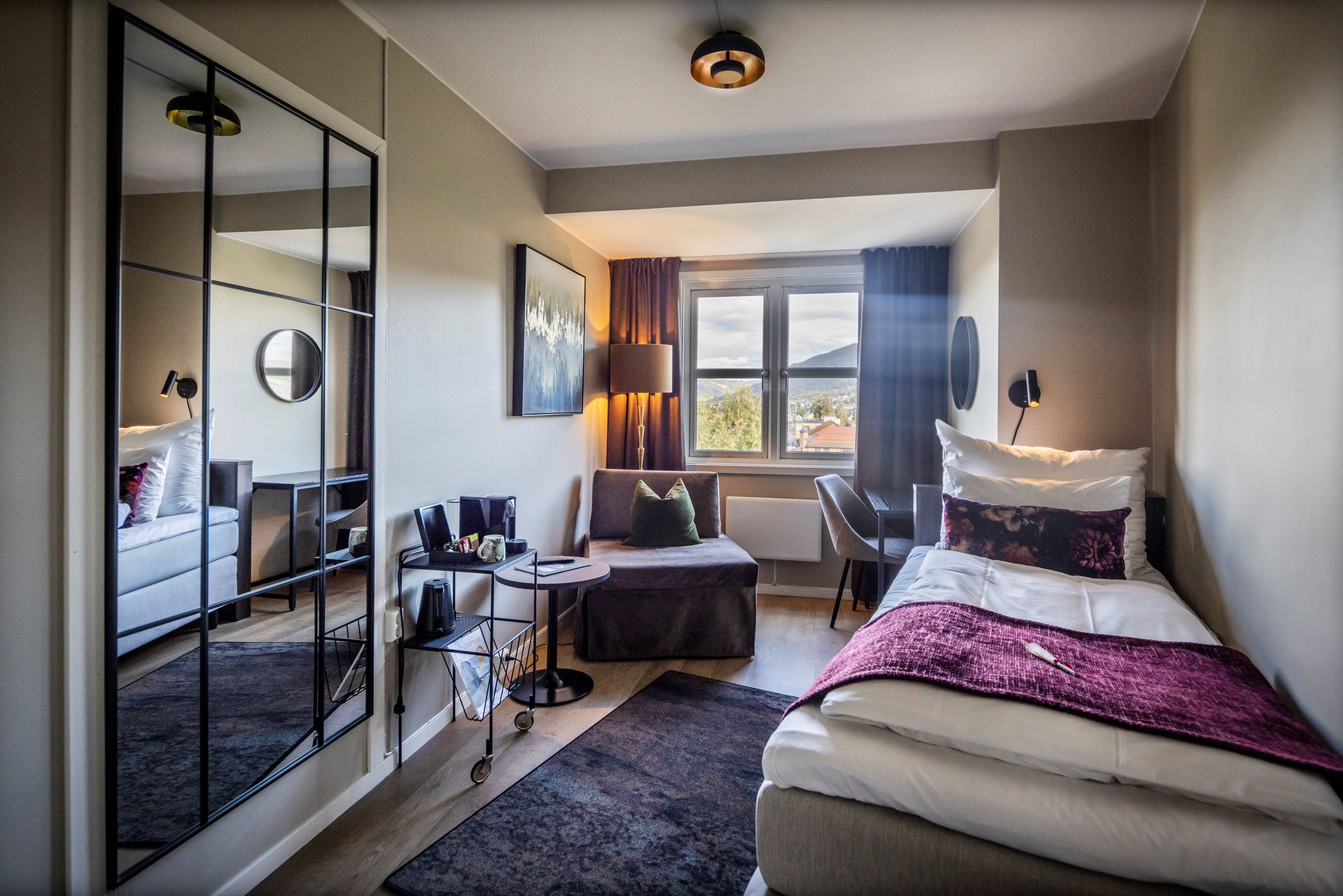 A hotel room with a bed, a desk, a chair, a mirror, a window with curtains, and a view of the outside.