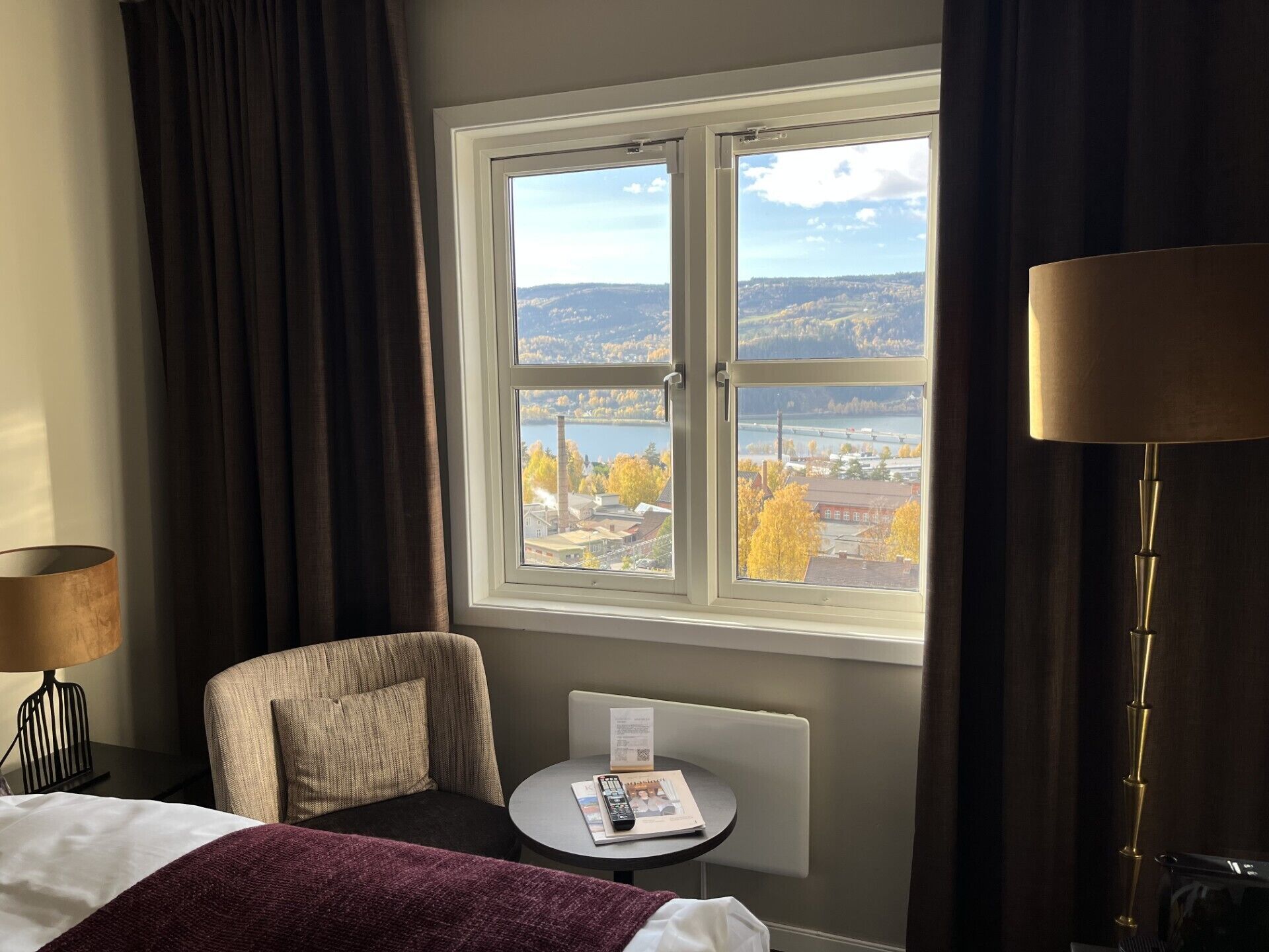 A hotel room with a bed, a chair, a small table with a remote control and a menu, and a window with a view of a city and hills.