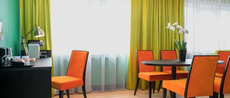 A modern hotel room with a wooden floor, orange chairs, a round table, and a desk with a lamp and a coffee machine.