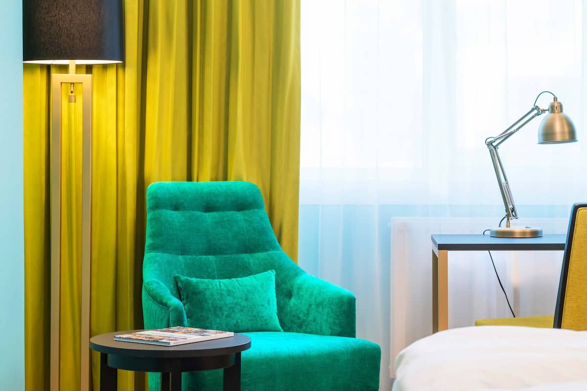 A hotel room with a green armchair, a small round table with a magazine, a lamp, and a bed with a yellow headboard.