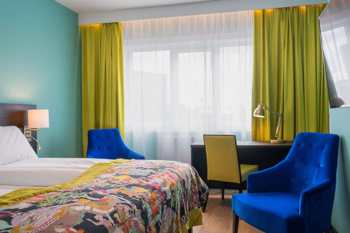 A hotel room with a bed, two blue armchairs, a desk with a chair, a lamp, and a window with sheer curtains.