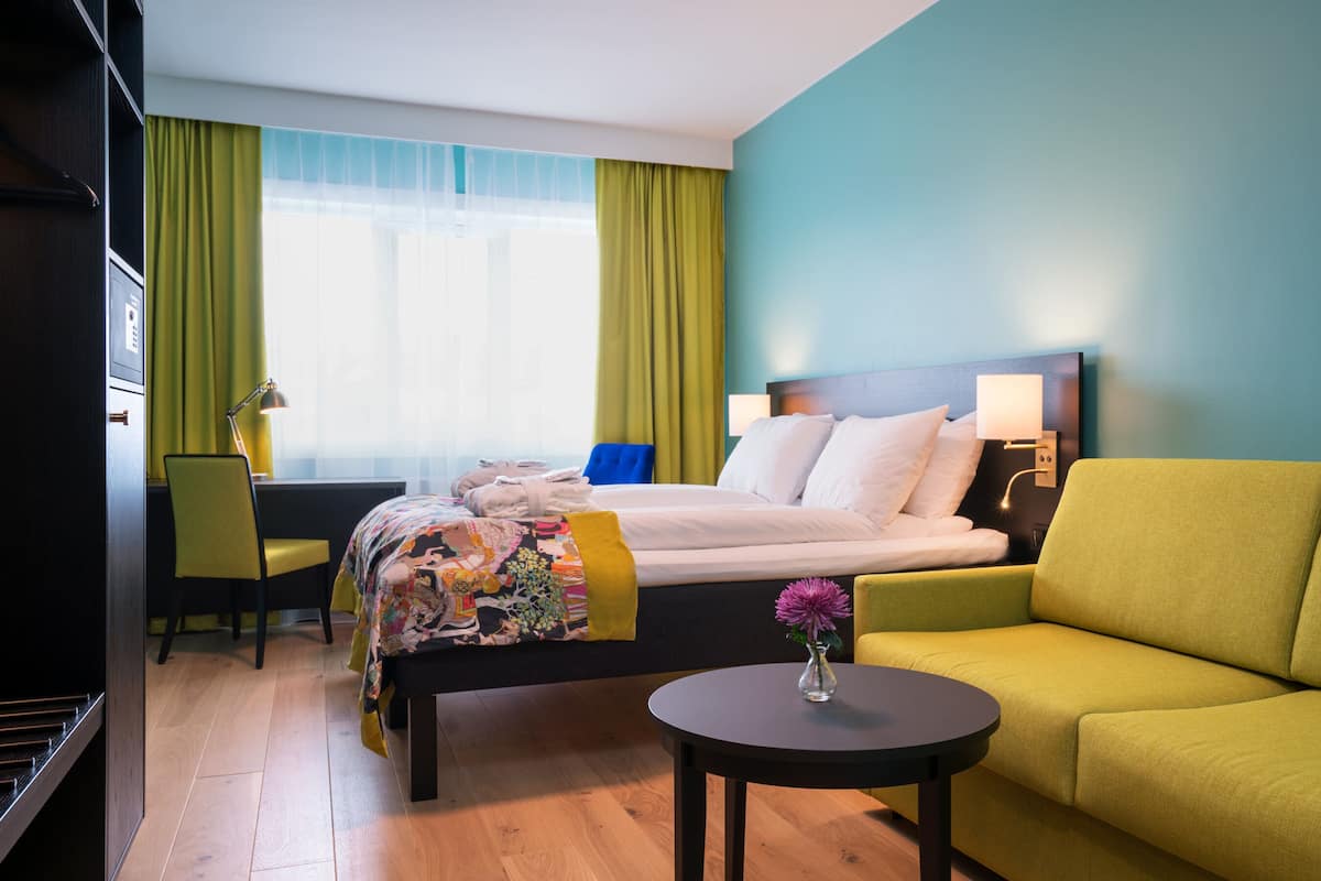 A hotel room with a bed, a green sofa, a desk with a chair, a small table with a vase, and a window with curtains.