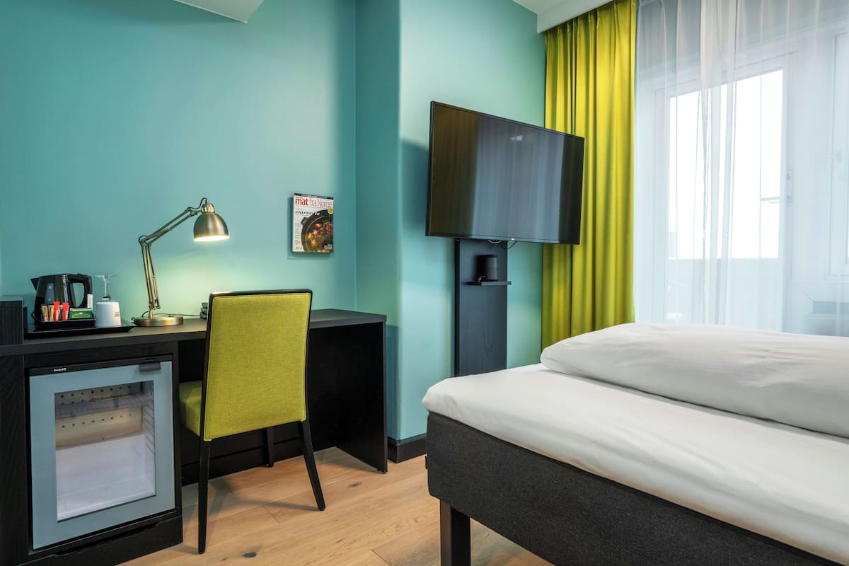 A hotel room with a bed, desk, chair, television, and a minibar.