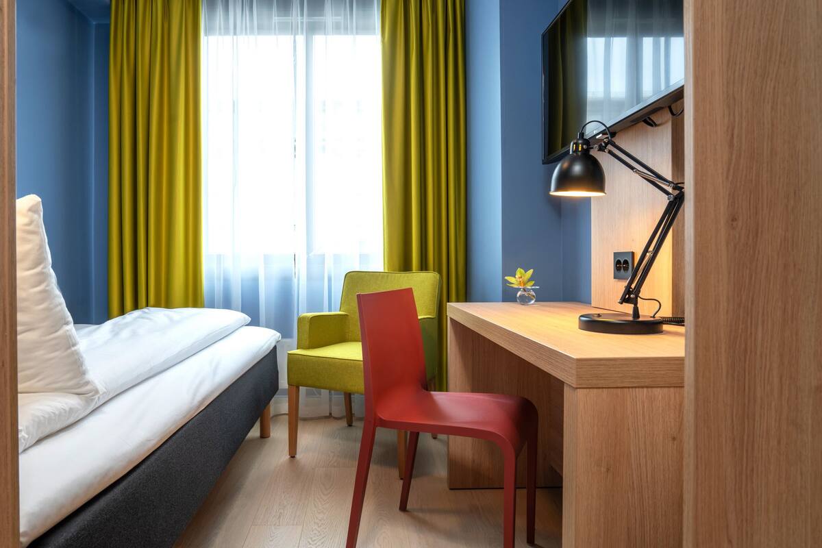 Une chambre d’hôtel avec un lit, un bureau, une chaise, une lampe de bureau et une fenêtre avec des rideaux.