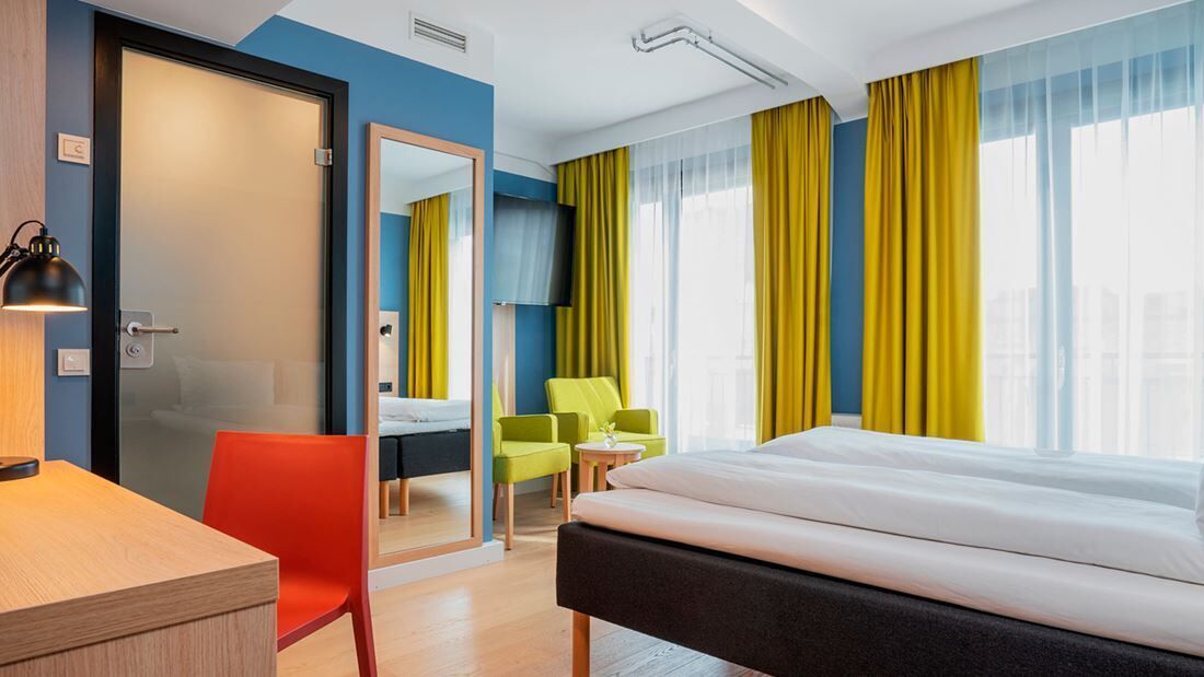 A hotel room with a bed, a desk, a chair, a mirror, and a window with curtains.