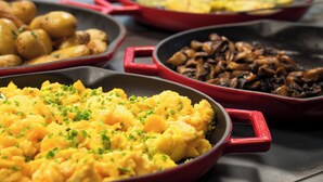Three red skillets on a stovetop, each containing different dishes: scrambled eggs, roasted potatoes, and sautéed mushrooms.