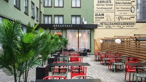 An outdoor cafe area with multiple tables and chairs, a sign for 'BREAKFAST', and a building facade with various signs.