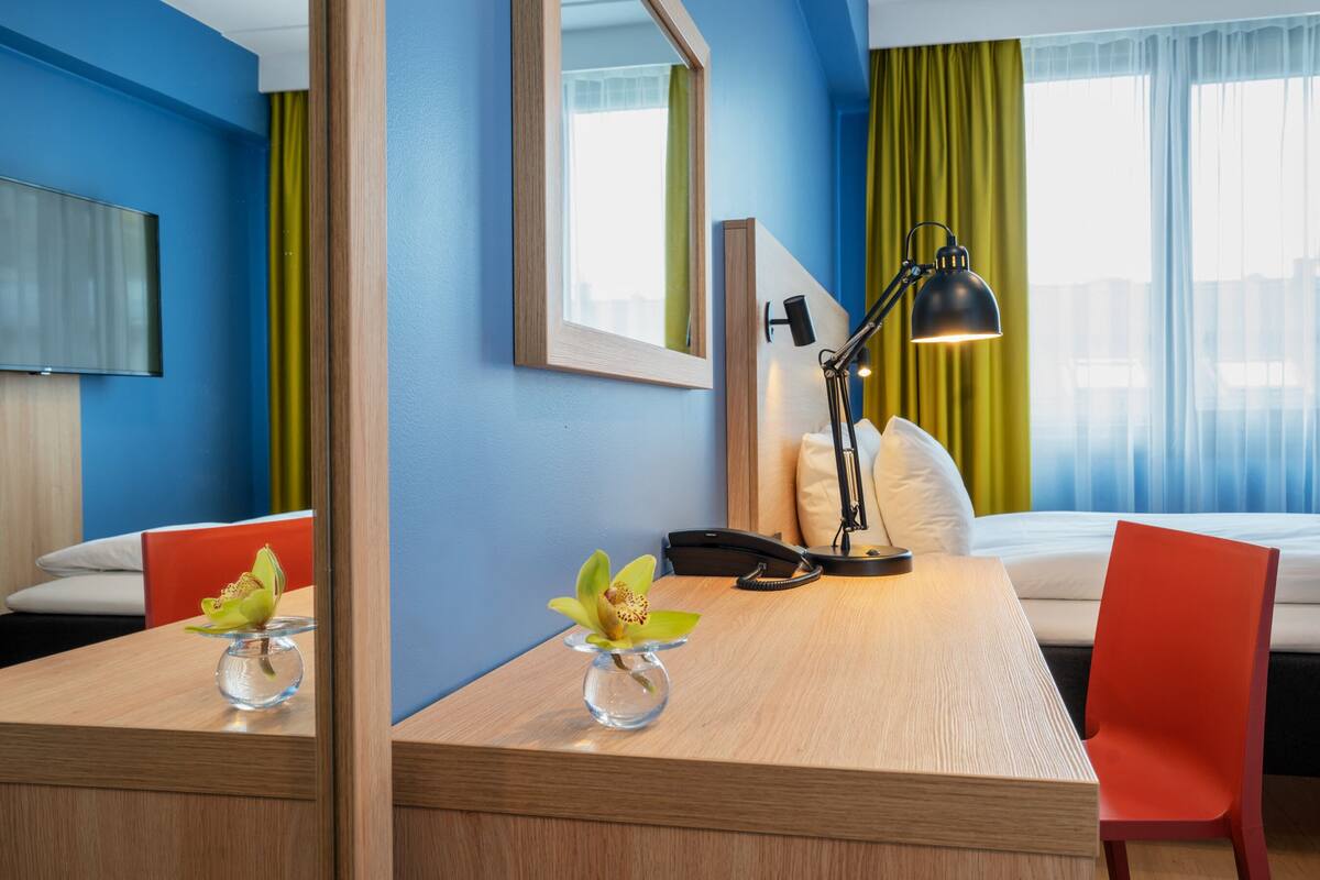 Une chambre d’hôtel avec un bureau en bois, une chaise, un lit, une lampe de chevet, un téléphone et un miroir.
