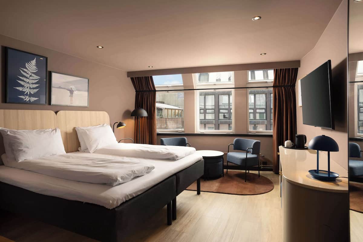 Una habitación de hotel moderna con una cama grande, dos sillas, una mesita y un televisor.