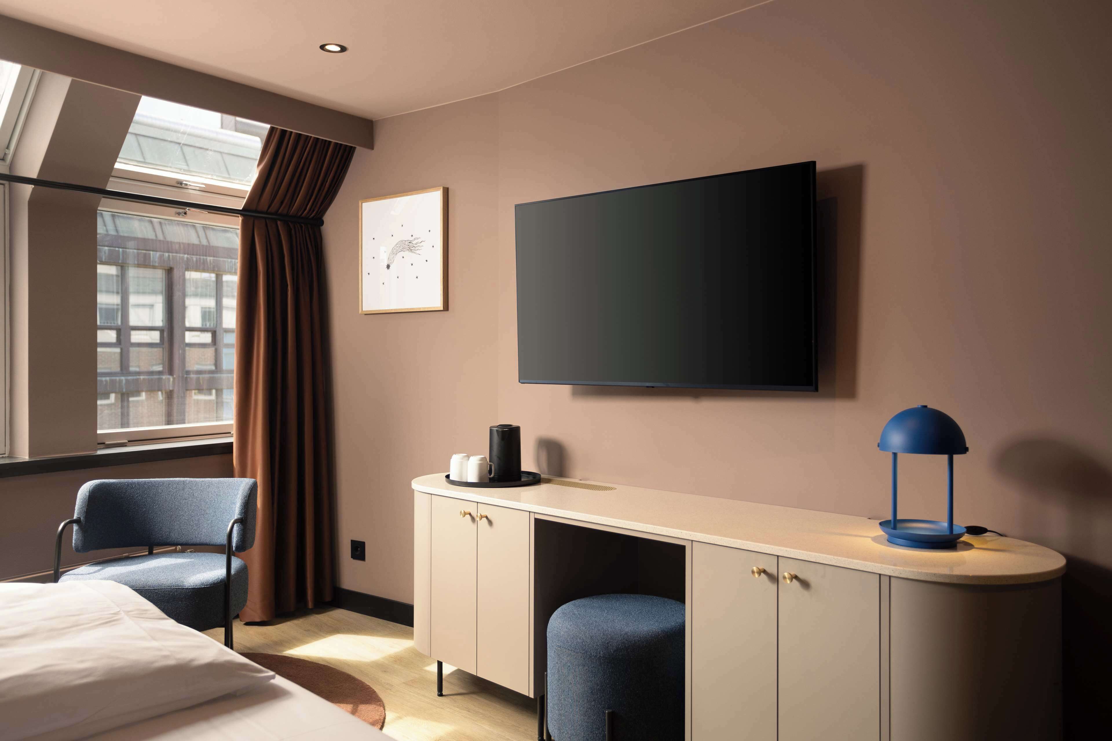 Et hotelværelse med en seng, to stole, et skrivebord med lampe, et fladskærms-tv og et vindue med gardiner.