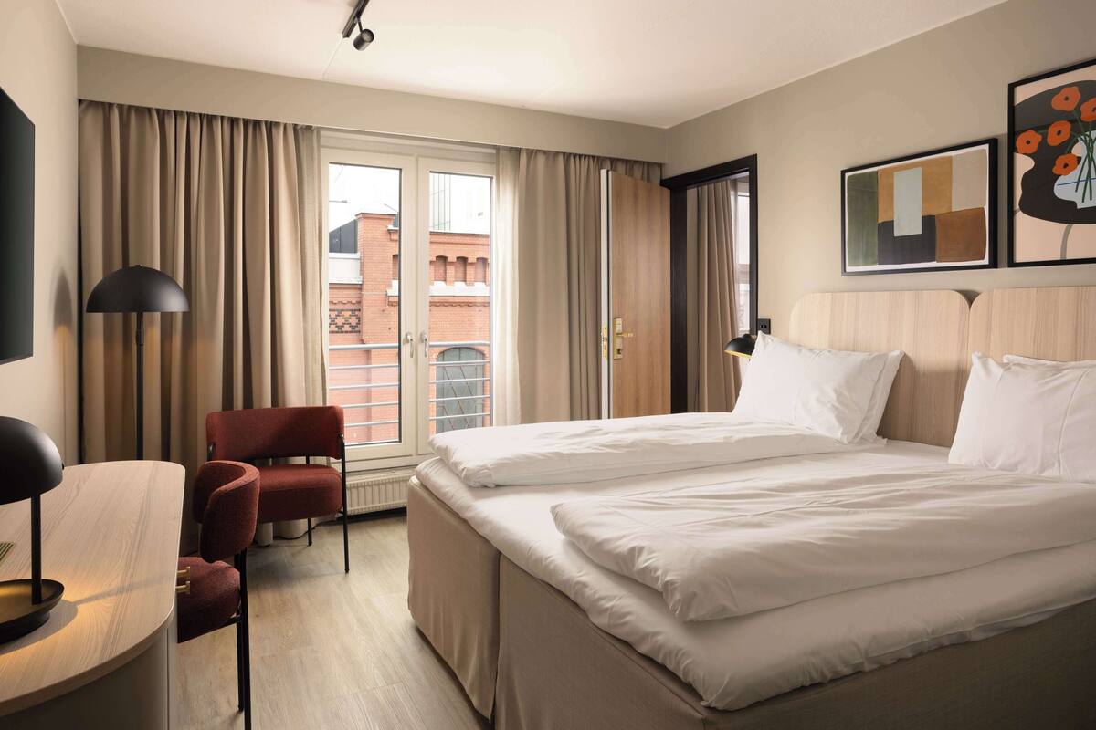 Una habitación de hotel con una cama, un escritorio, una silla y un ventanal con cortinas.