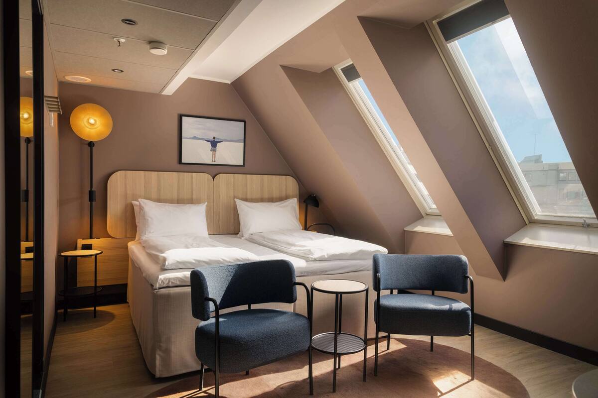 Una habitación de hotel moderna con una cama, dos sillas, una mesita, una lámpara de pie y un cuadro en la pared.
