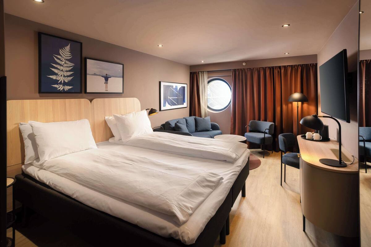 Habitación de hotel con una cama grande, un escritorio con lámpara, dos sillas, un ventanal redondo y obras de arte en las paredes.
