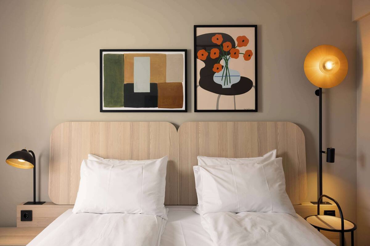 Una cama doble con sábanas blancas, un cabecero de madera, una lámpara de pie negra y dos cuadros en la pared.