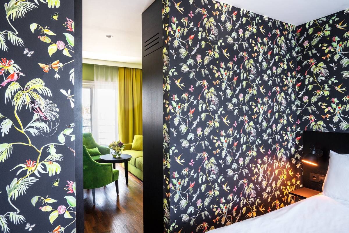 A hotel room with a floral wallpaper, a green armchair, a small table with a vase, and a bed with a white headboard.