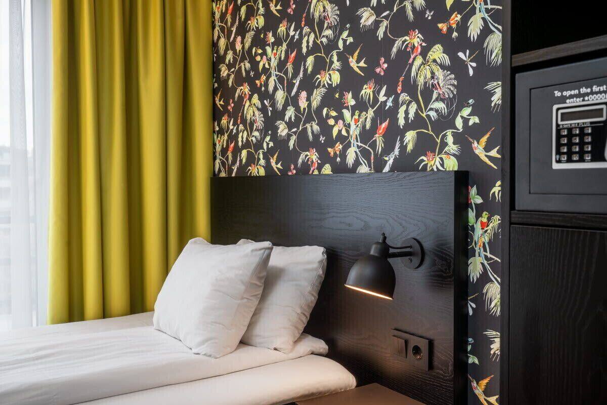 A hotel room with a bed, a nightstand, a wall-mounted lamp, and a door with a keypad lock.