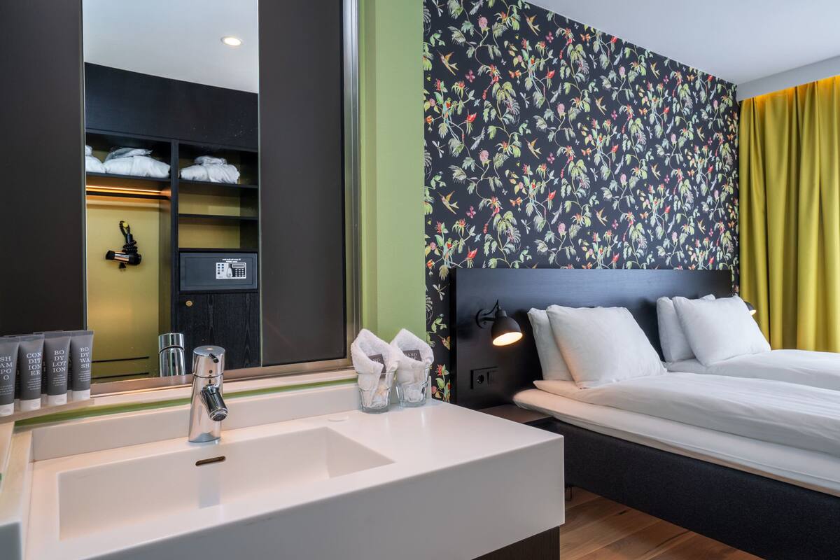 A hotel room with a bed, a bedside table with toiletries, a mirror, and a wall with a floral pattern.