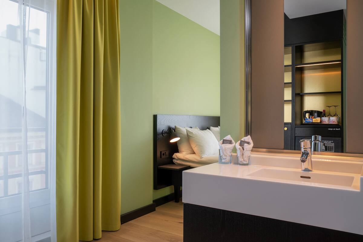 A modern hotel room with a large window, a bed with white linens, a bedside table with a lamp, and a built-in minibar.