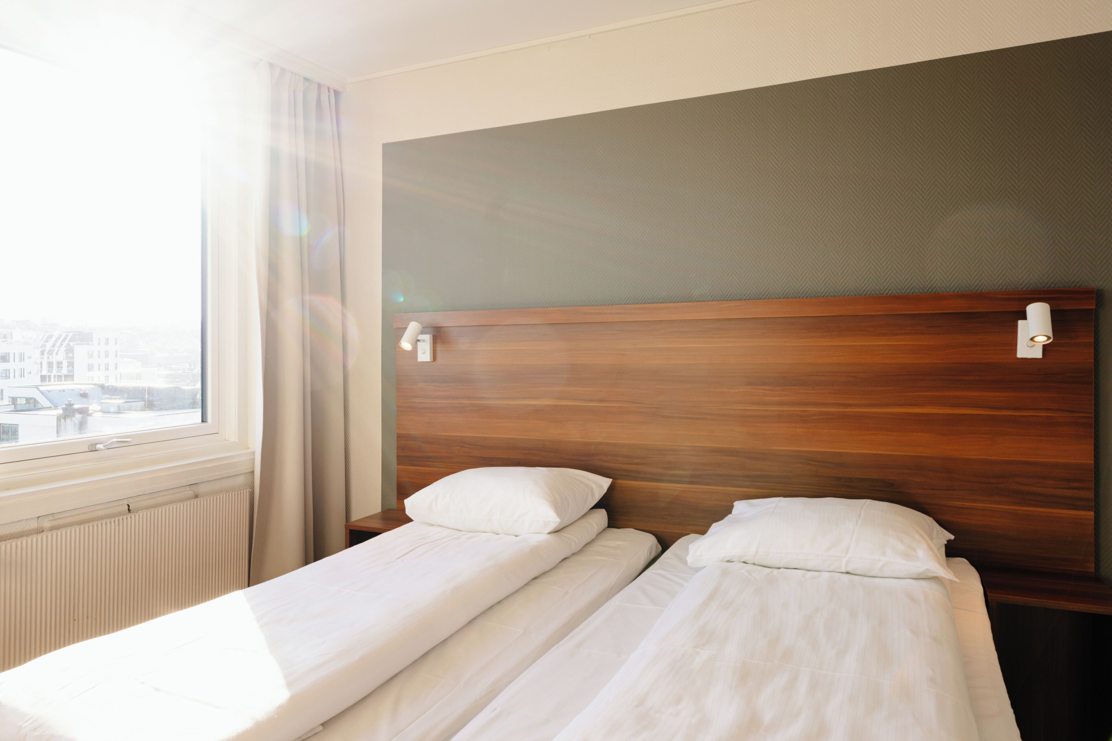Une chambre d’hôtel avec deux lits simples, une tête de lit en bois, une fenêtre avec des rideaux et une vue sur les bâtiments environnants.
