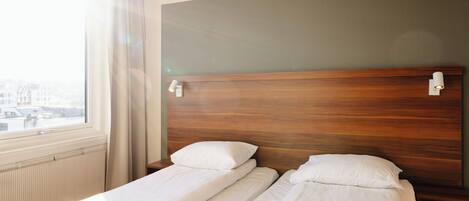 A hotel room with two single beds, a wooden headboard, a window with curtains, and a view of buildings outside.
