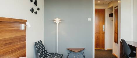 A hotel room with a bed, a desk, a chair, a small table, and a floor lamp.