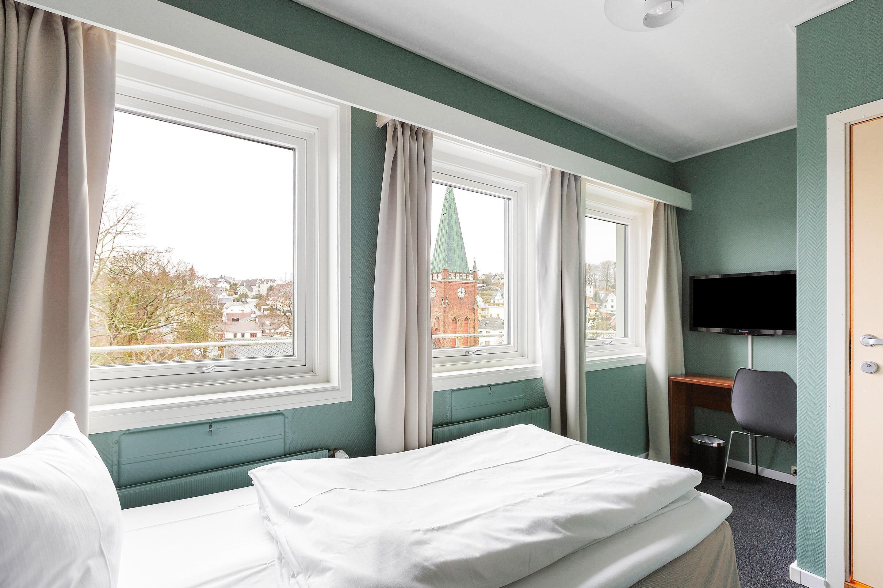 A hotel room with a bed, a desk, a chair, a television, and large windows with curtains.