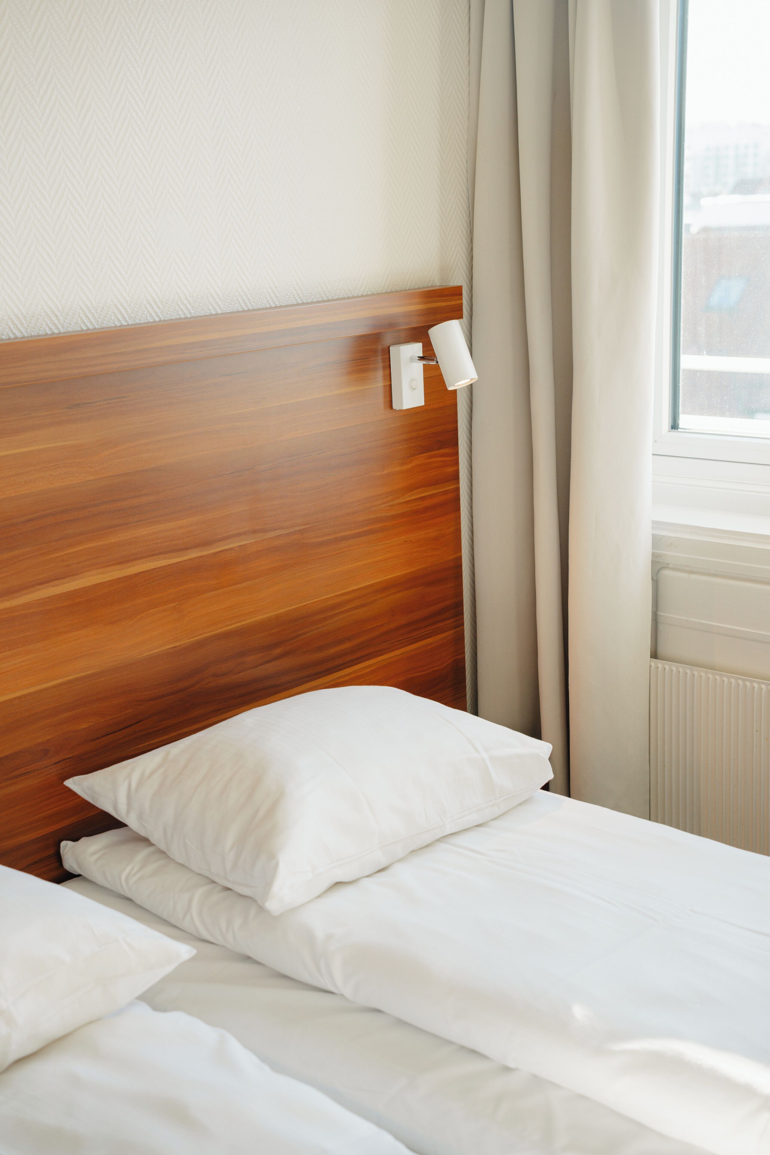 A neatly made bed with white linens and pillows, a wooden headboard, and a window with curtains.