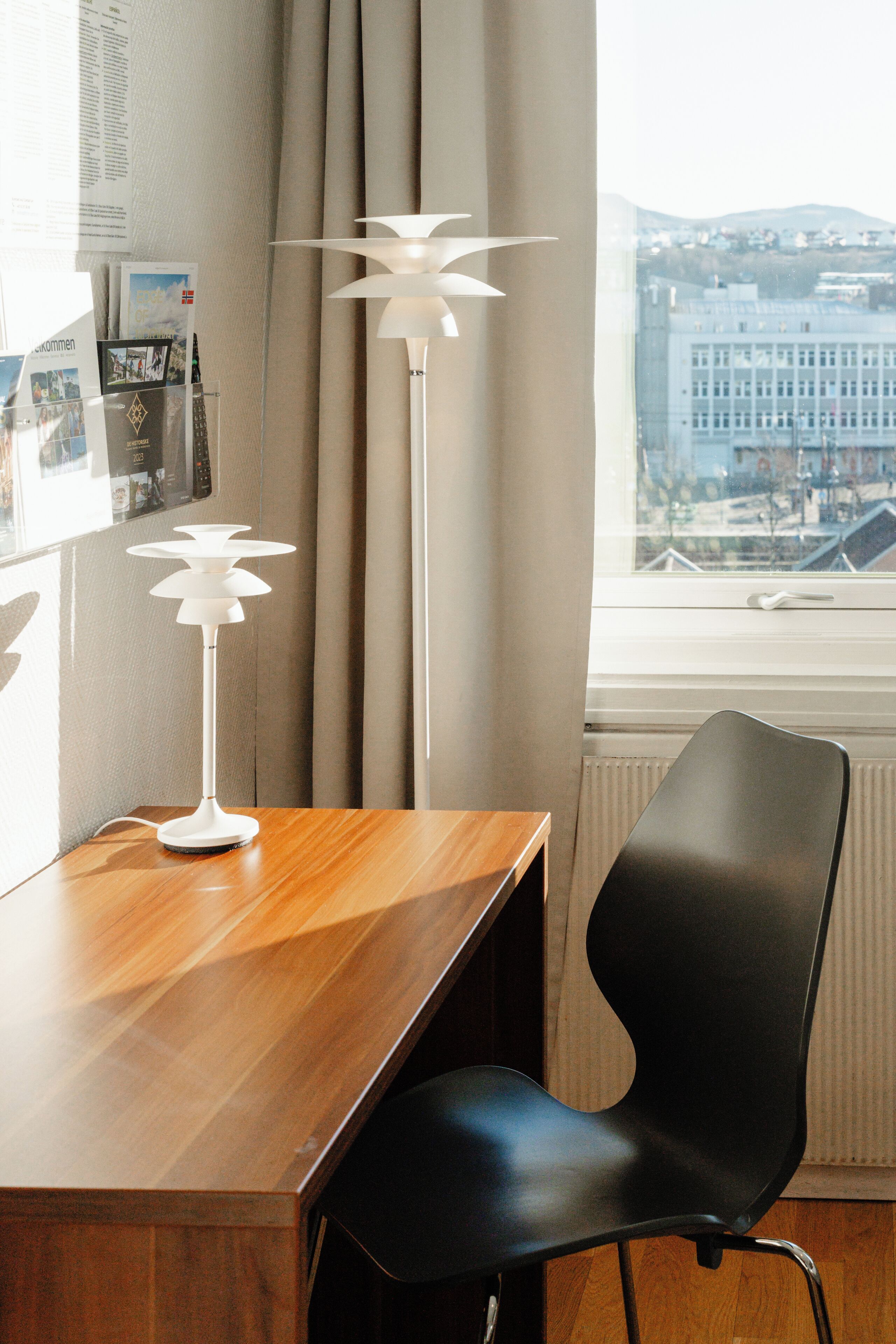 Una habitación moderna con un escritorio de madera, una silla negra, una lámpara de pie blanca y vistas a un paisaje urbano a través de una ventana con cortinas.