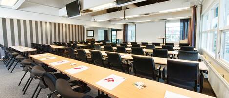 Un salón de conferencias con varias mesas y sillas, una pantalla de proyector y ventanas amplias.