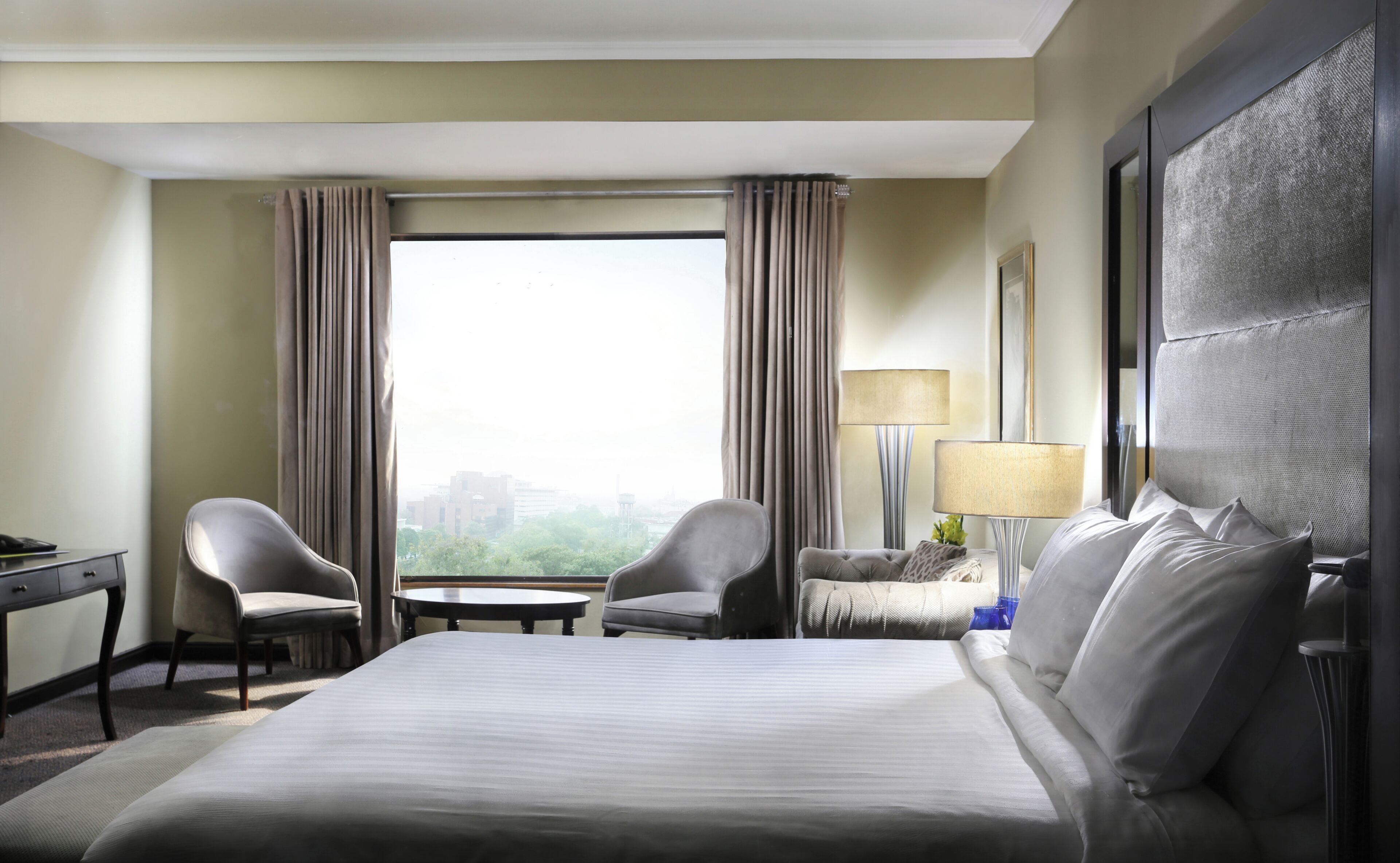 A hotel room with a bed, two chairs, a desk, a lamp, and a large window with curtains.