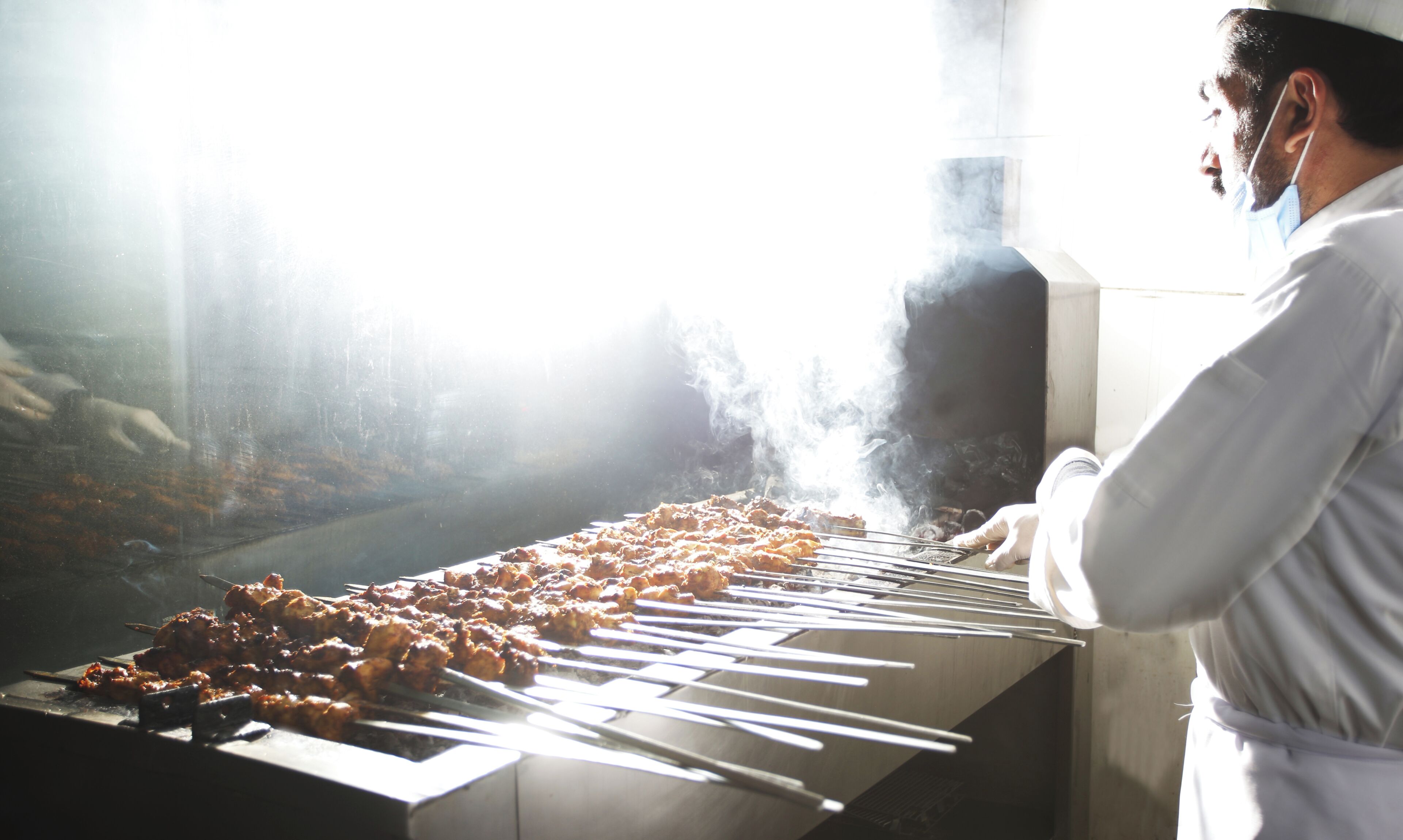 A person grilling skewers on a metal grill with a view of a building in the background.
