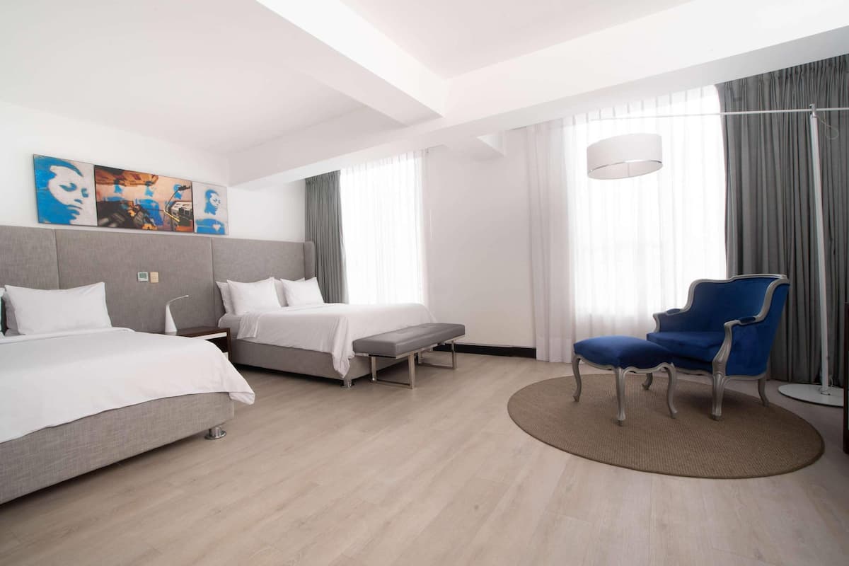 A hotel room with two beds, a bench, a blue armchair, and a large window with curtains.