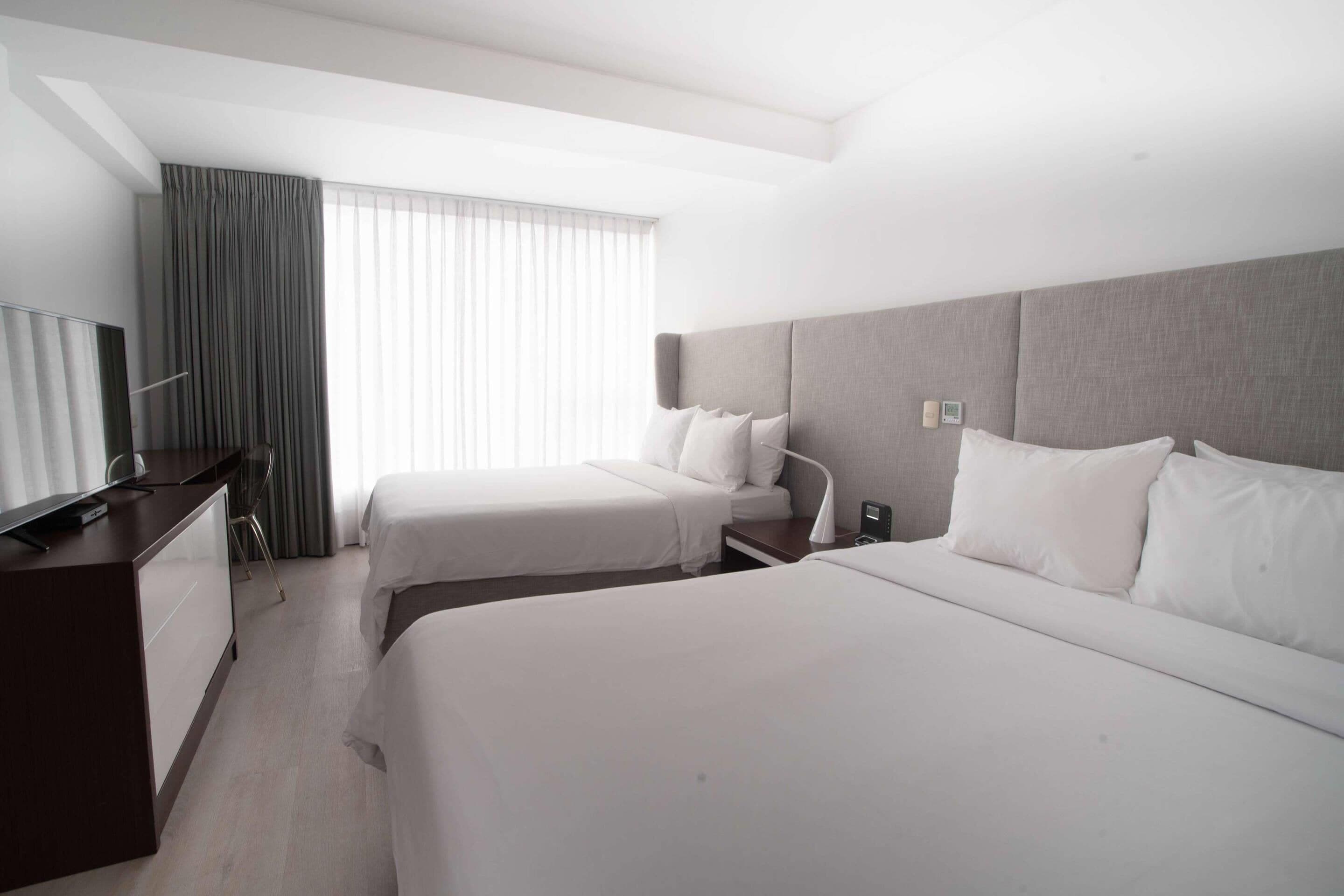 A hotel room with two beds, a desk, a TV, and a window with curtains.