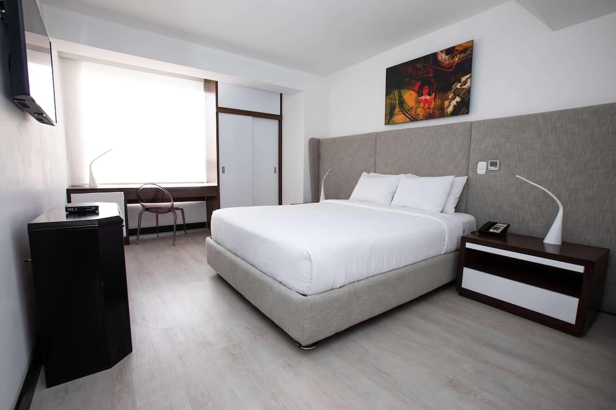 A modern hotel room with a large bed, a desk, a chair, and a painting on the wall.
