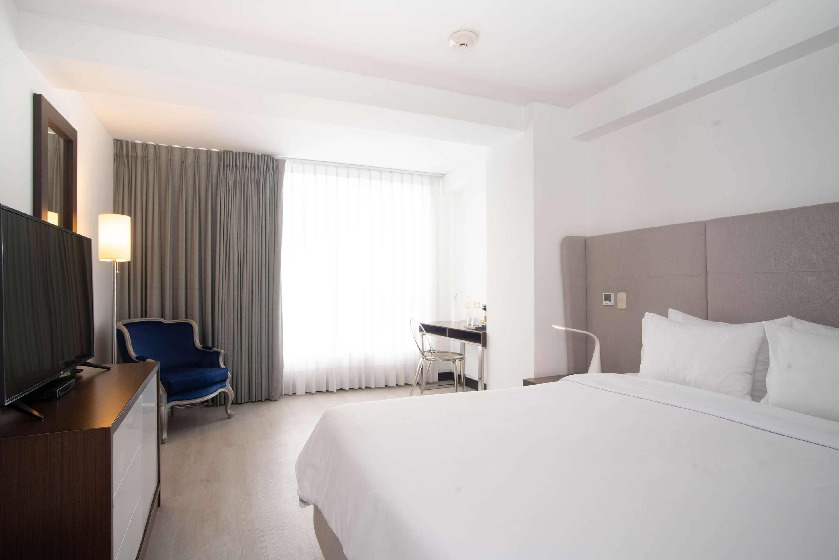 Una habitación de hotel con una cama grande, un escritorio, una silla, un televisor y una ventana con cortinas.