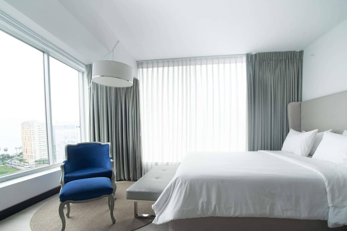 A hotel room with a large bed, a blue chair, a round table, and a view of the city through large windows.