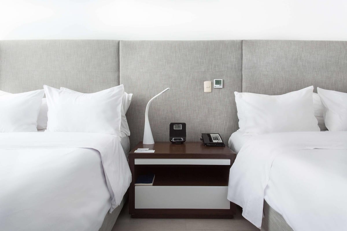 A hotel room with two beds, a nightstand with a lamp, a phone, and a small shelf.