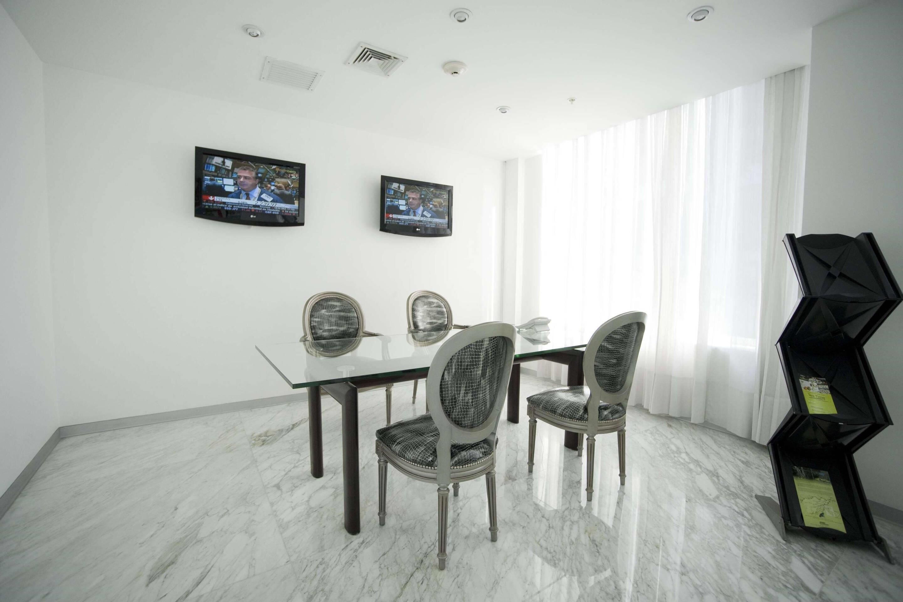 A dining area with a glass table, four upholstered chairs, two wall-mounted televisions, and a black geometric bookshelf.