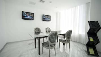 A dining area with a glass table, four upholstered chairs, two wall-mounted televisions, and a black geometric bookshelf.