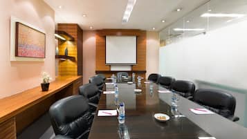 A conference room with a long table, black leather chairs, a projector screen, and a wooden desk with a plant.