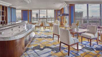 A hotel lobby with a marble reception desk, seating area with chairs and tables, and large windows offering a city view.