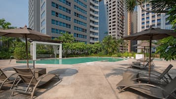 A poolside area with lounge chairs and umbrellas, surrounded by tall buildings and greenery.