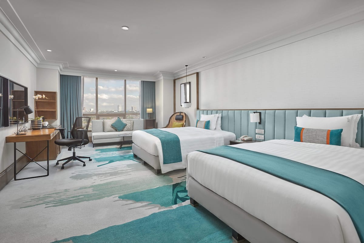 A hotel room with two beds, a desk, a chair, a TV, and a large window with a city view.