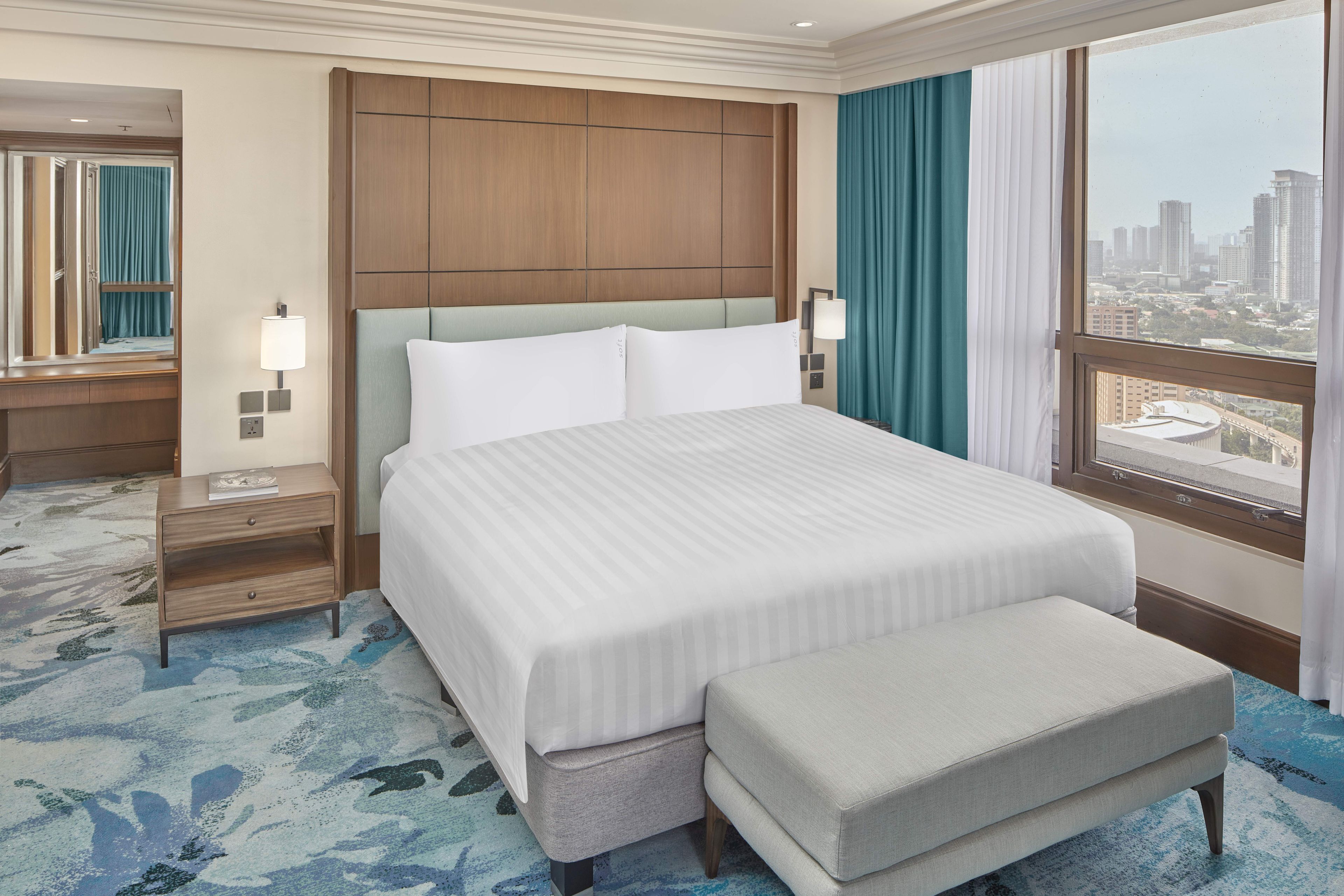 A hotel room with a large bed, a nightstand, a mirror, a window with a city view, and a blue patterned carpet.