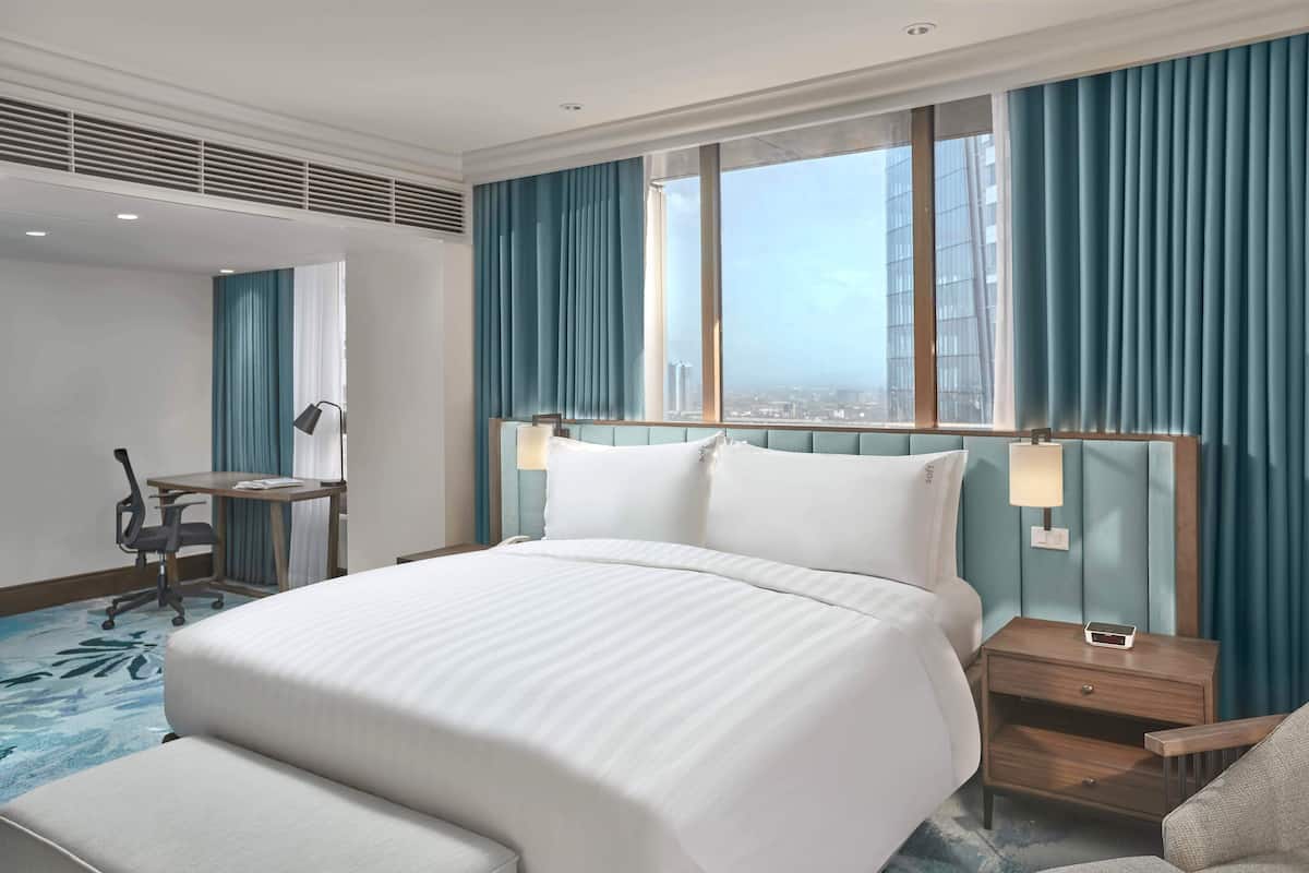 A hotel room with a large bed, a desk, a chair, and a view of the city through the window.