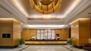 Ein Hotel-Empfangsbereich mit modernem Design, der ein zentrales Lichtelement, einen Empfangstresen und Kübelpflanzen zeigt.