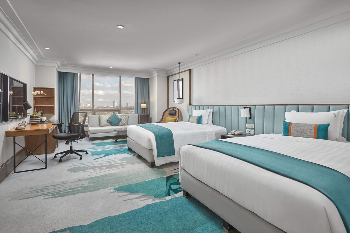 Une chambre d’hôtel avec deux lits, un bureau, une chaise, une télévision et une grande fenêtre donnant sur la ville.