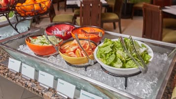 A buffet with labeled bowls of various vegetables like cucumber, onion, tomato, and romaine lettuce.